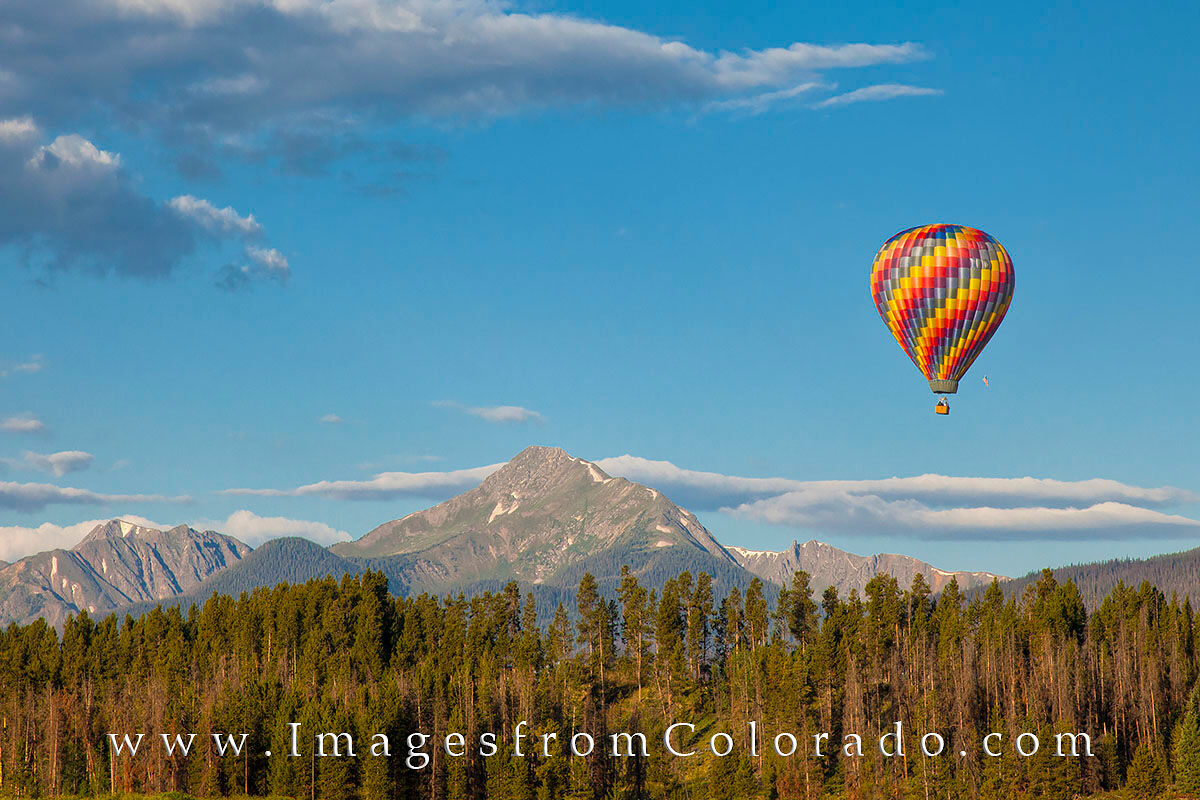 Hot Air Baloon over Fraser, Colorado 1 On a cold summer morning, a hot air baloon drifts over the pine trees near Fraser, Colorado. In the distance is the locally well...